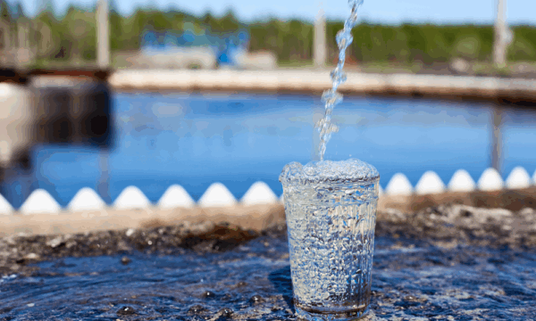 A clear glass overflowing with water, positioned on a wet surface outdoors, with a stream of water pouring into it. In the blurred background, there is a large body of water and green trees under a blue sky.