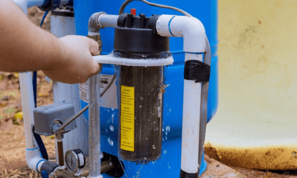 A hand uses a wrench to loosen the housing of a water filter, causing water to leak from the unit.